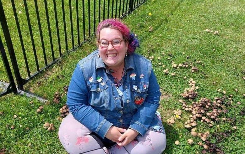 A woman with pink hair in a pony tail is seated on a grassy area, smiling up at the camera. She is wearing glasses, a denim jacket and purple leggings.