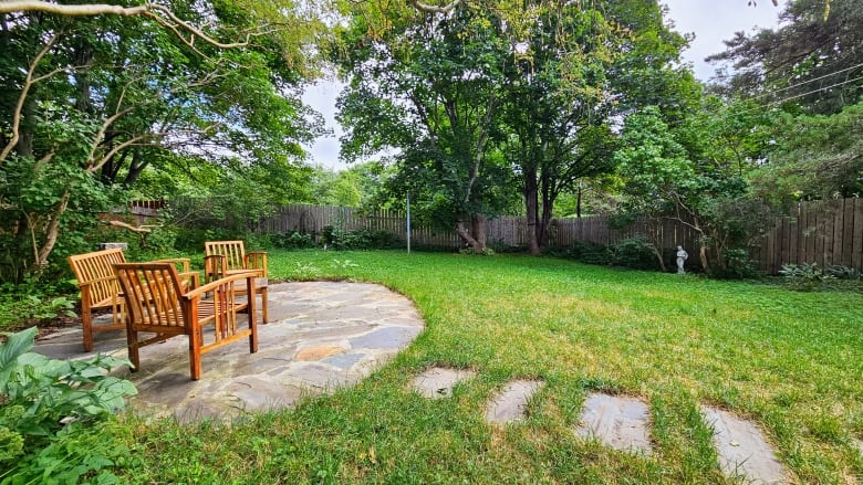 Two wooden chairs and a wooden bench are arranged on a circular space inlaid with stone, sitting in a fenced backyard, overlooked by trees.