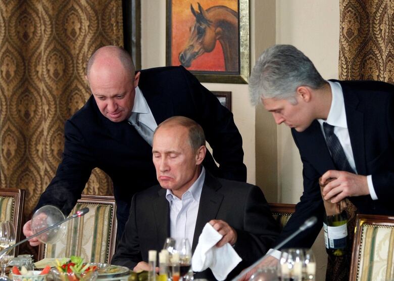 Two men hover over another man who is eating what appears to be a fine dining meal.