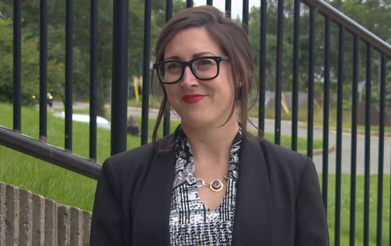 A woman is wearing red lipstick and black glasses. She is standing outdoors, in front of a black railing. 