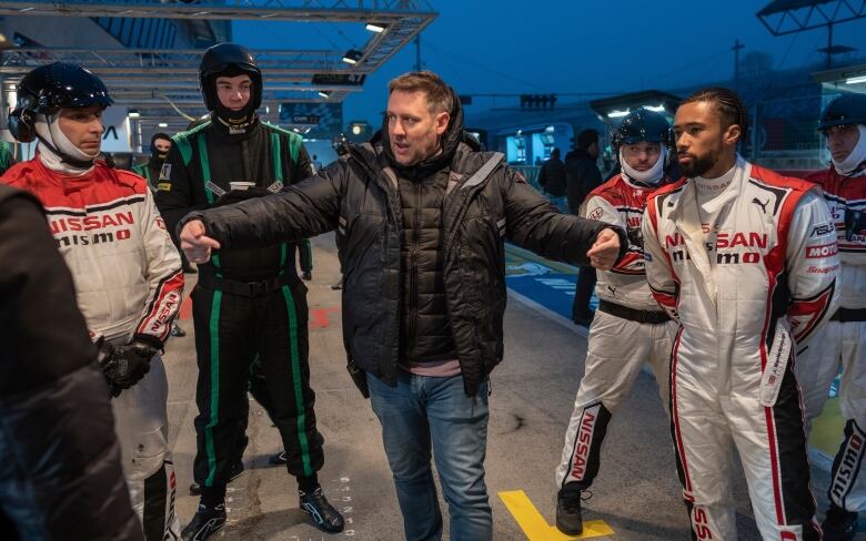 A man in a jacket and jeans stands amid other men wearing race gear on an outdoor race track.