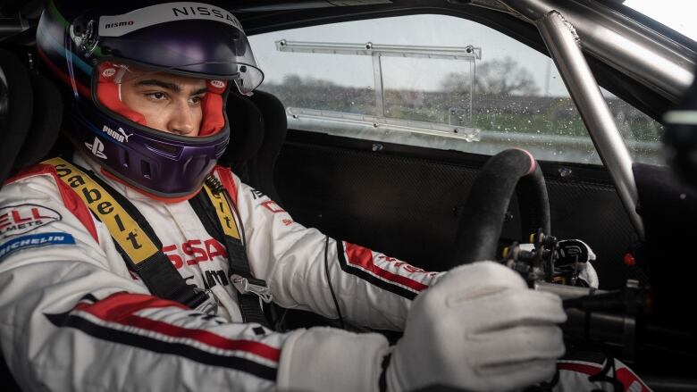 A man wearing race gear and full visor helmet grips the wheel of a race car. 