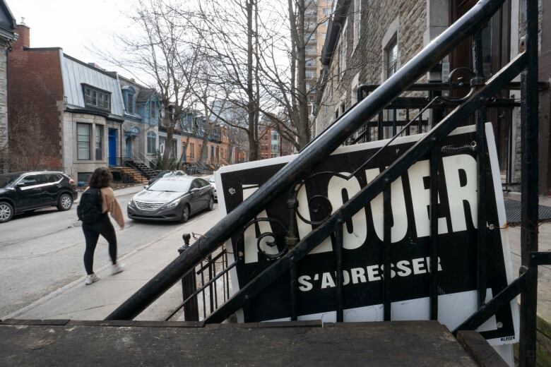 A sign that says for rent in French, on a staircase railing. A young woman walks on the street nearby.