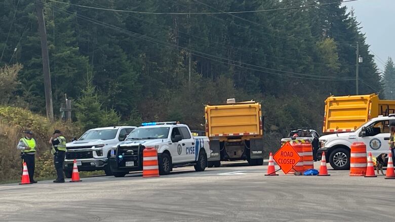 Two people in high visibility vests stand beside several vehicles blocking a highway.