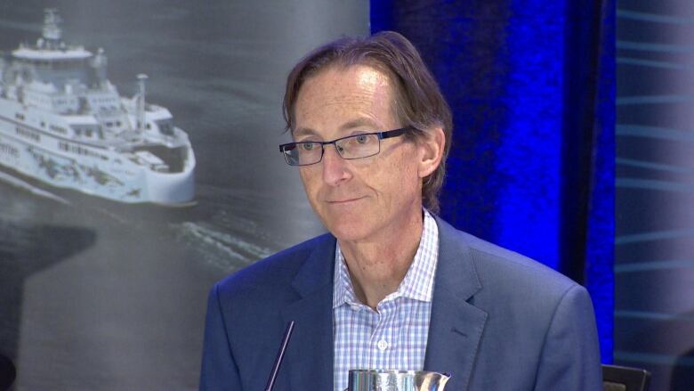 A man in a suit and open-collar white and blue striped shirt sits in front of a picture of a ferry with a blue curtain behind him.