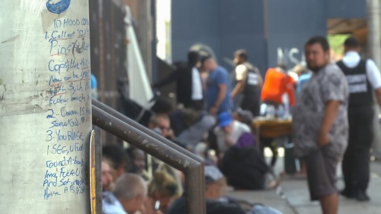 A crowd of people mill about on the sidewalk. In the foreground graffiti details the 