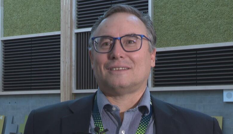 A man smiles at the camera. He has glasses on and is wearing a suit. 