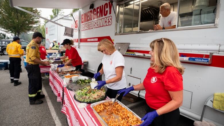 Firefighters line up at a long table where people smile and serve food in front of a large food trailer that says, Disaster Emergency Services.