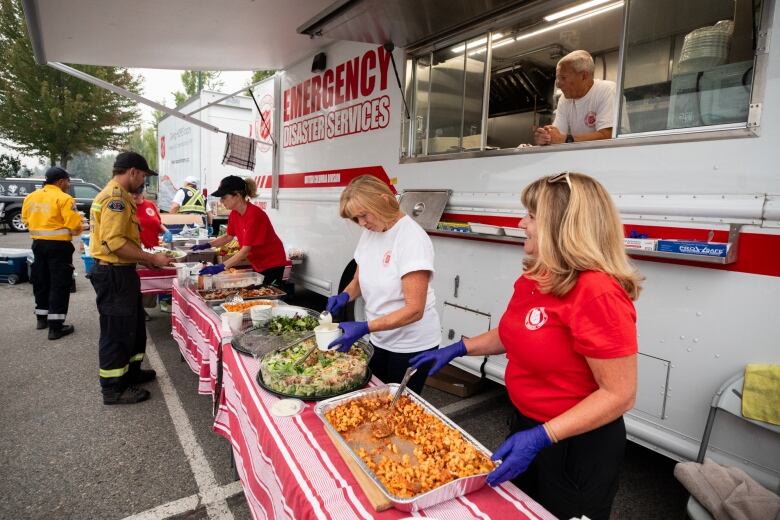 Firefighters line up at a long table where people smile and serve food in front of a large food trailer that says, Disaster Emergency Services.