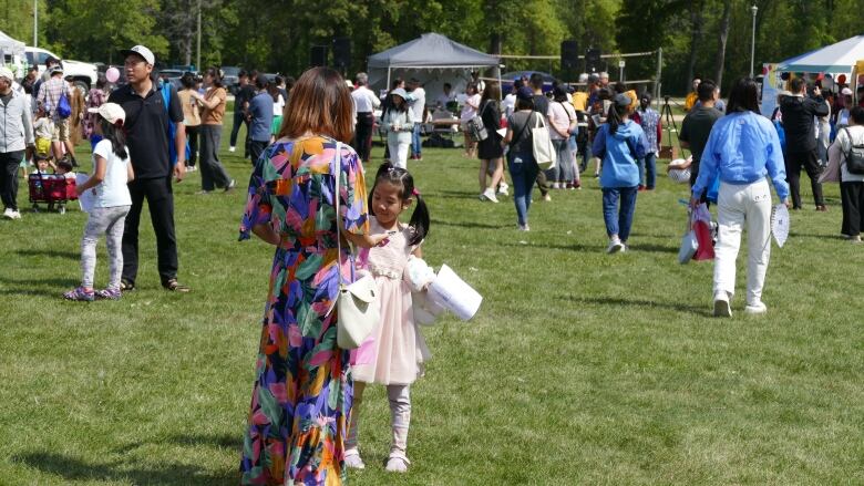 A woman and a young girl stand in a field. A crowd of other people walk in the background.