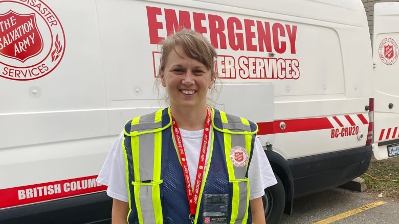 A blonde white woman wearing a high-vis jacket poses in front of a white van.