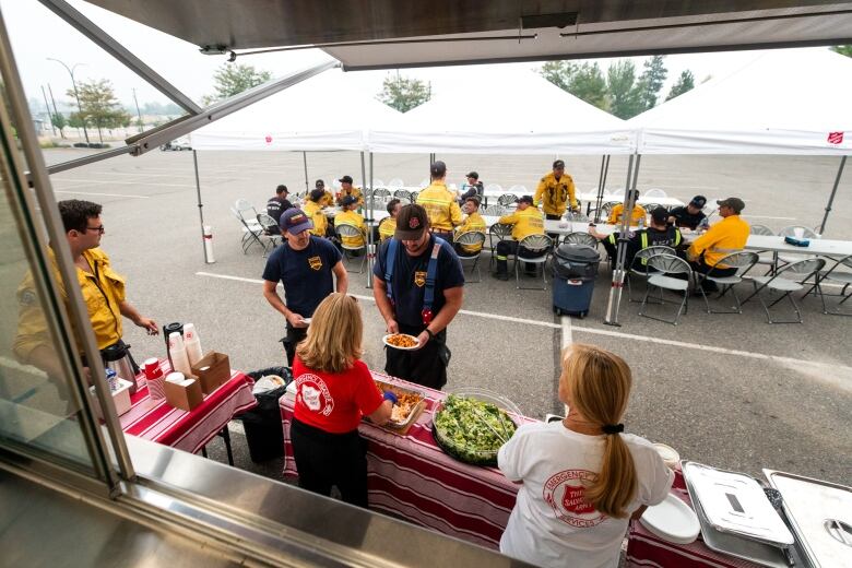 Two men queue up at a food service table, with a dozen other men behind them wearing yellow.