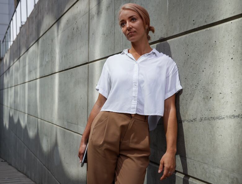 A woman with pinkish blonde hair leans against a stone wall. She is wearing a button up shirt and high waisted pants. 