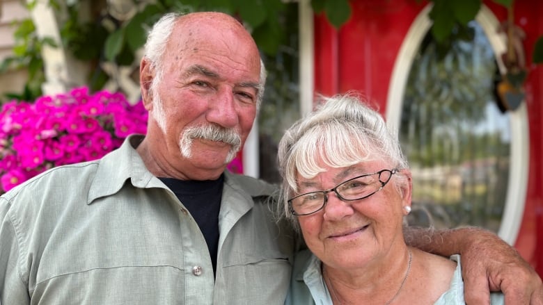 A couple in their 70s stand in front of a red door. 