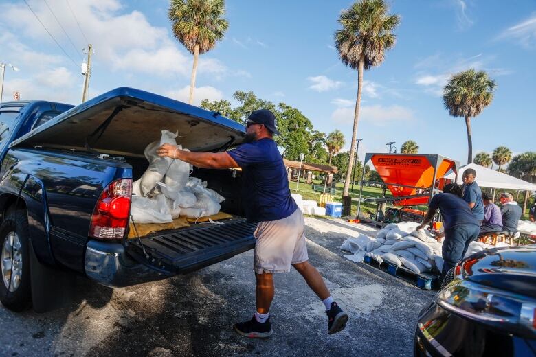 A man wearing a blue T-shirt and backwards hat throws sandbags into the bed of a dark blue pickup truck.