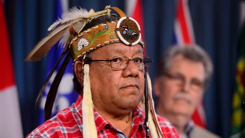 A man wearing a traditional Indigenous headdress speaks at a press conference.