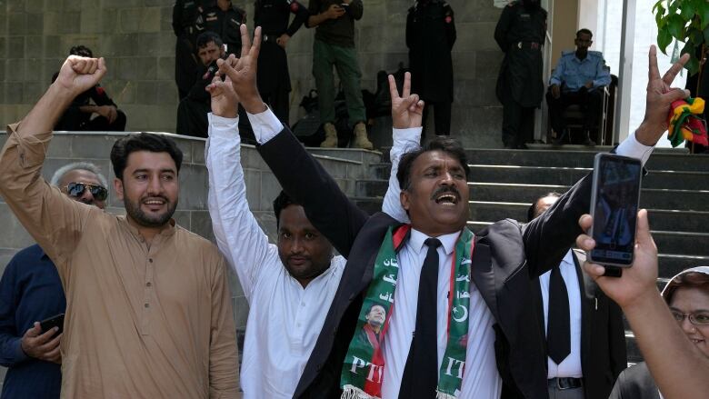 Three men are shown lifting their arms in exultation while standing outside.