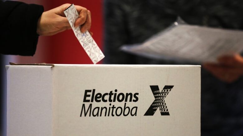 A hand holds a ballot above a ballot box.