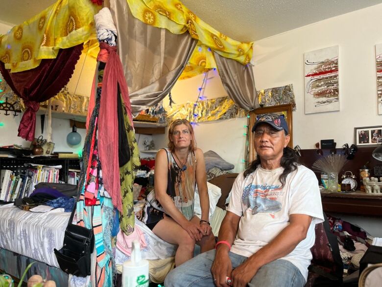 A woman with short blond hair sits on a bed next to a man with long black hair, wearing a ball cap.