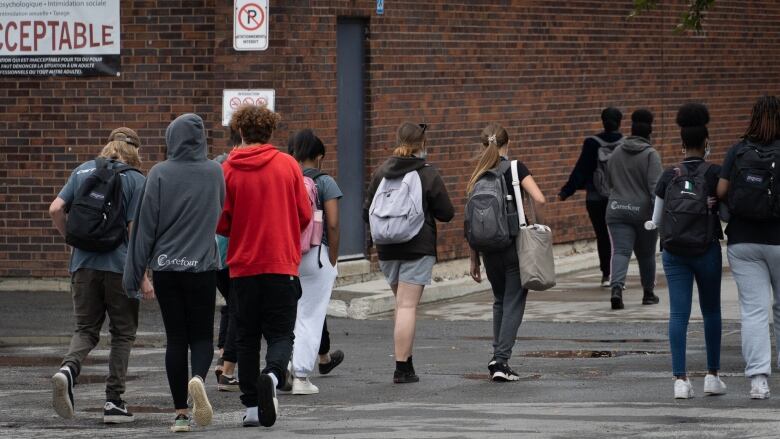 Seen from behind, several teenagers with book bags and backpacks walk toward a brick building.