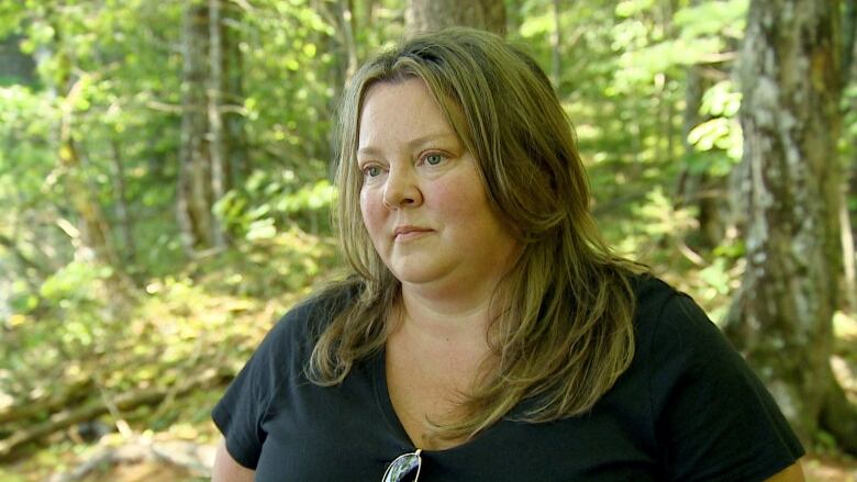 A white woman in a black t-shirt and long, dark blonde hair stands in a green wooded area