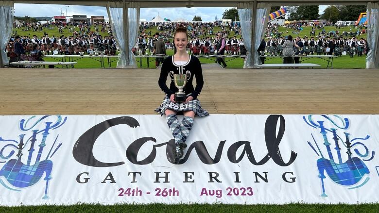 A highland dancer is seated on the stage holding her trophy after winning a world championship. 