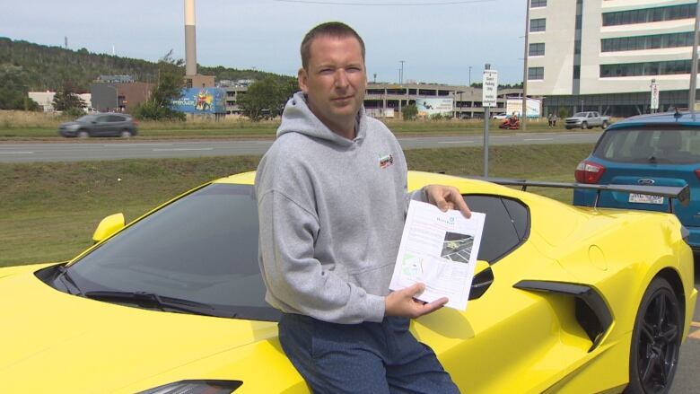 A man is leaned against his yellow Corvette, with a printed speed detection notice in his hand. 