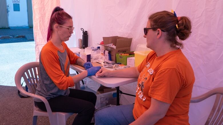 Two people wearing orange shirts sit on chairs beneath a tent.