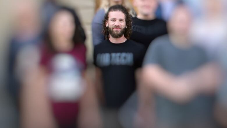 A man stands in a group of blurred out people. He has long curly hair and is wearing a t-shirt.