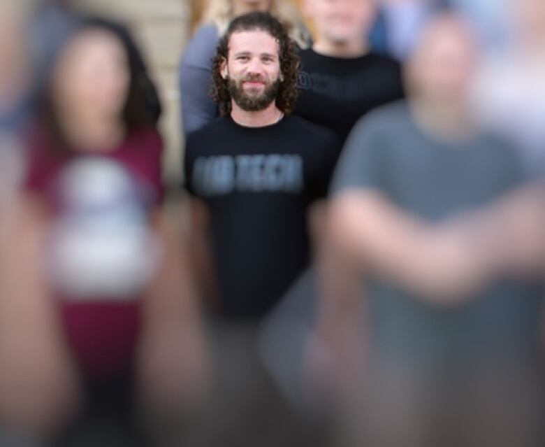 A man stands in a group of blurred out people. He has long curly hair and is wearing a t-shirt.