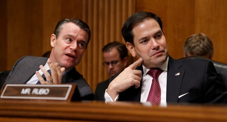 Two men in suits sit at a conference table.