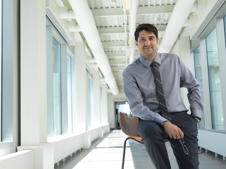 A man in a grey shirt, dark pants and a striped tie is seen smiling at the camera.