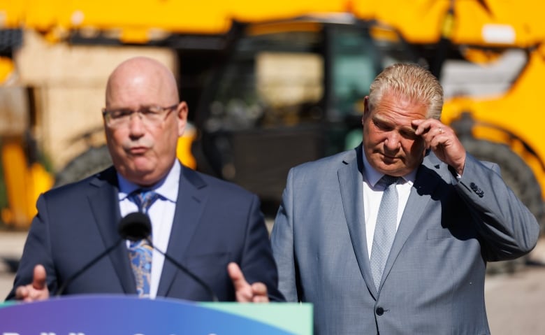 A man in a suit speaks at a lectern while another, standing to one side, puts his hand to his forehead.  