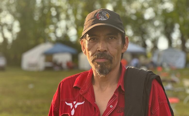 A man with a serious expression in a baseball hat, wearing a red button-up shirt looks at the camera. It's late afternoon, and the sunlight hits the side of his face.