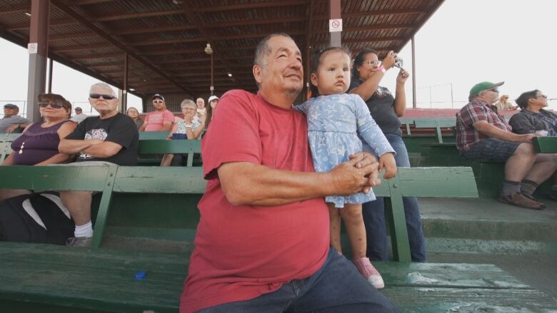 A man in a red T-shirt holds a toddler with pigtails next to him on a wooden bench. They're in a grandstand, watching a horse race.