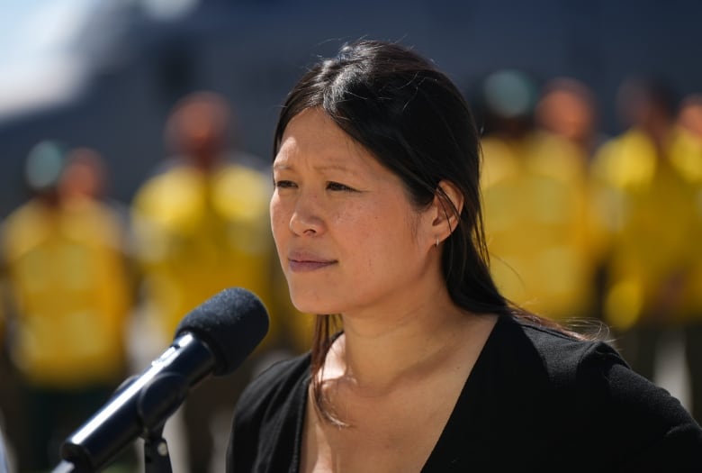 An East Asian woman is pictured at an outdoor news conference, with firefighters wearing yellow visible in the background.