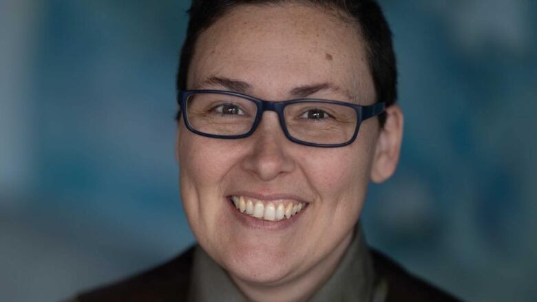 A person with cropped dark brown hair and rectangular glasses, wearing a black vest over a grey button-up shirt and blue patterned tye, smiles brightly for the camera.