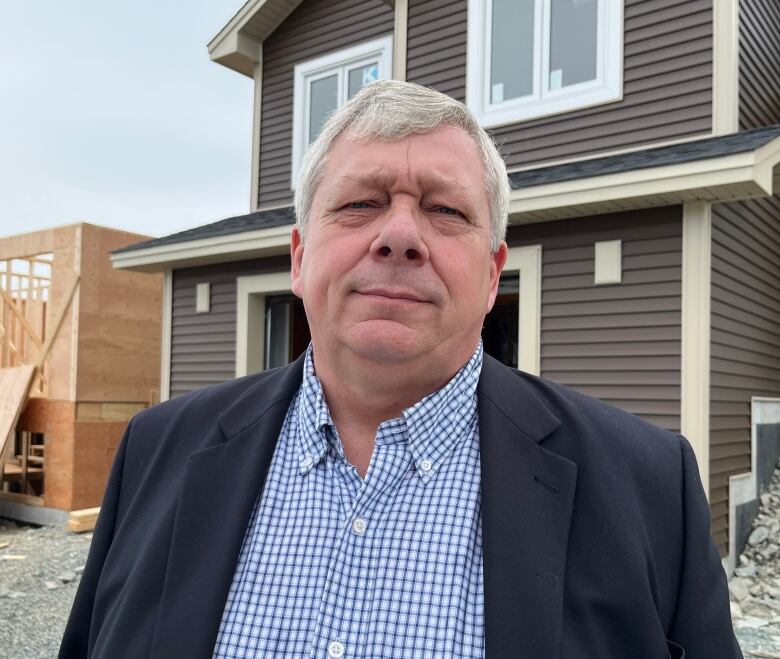 A man looks straight ahead. Behind him are two houses, both in different construction stages.