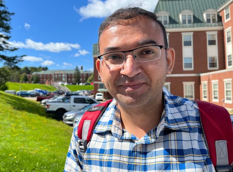 A man with glasses and a blue plaid shirt wearing a red backpack. He is standing in front of a parking lot and a red brick building.