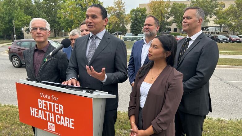 A man speaks at a podium, flanked by several NDP candidates. 