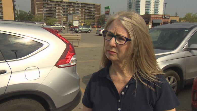 A woman with blond hair and glasses with a dark frame speaks with a reporter in a parking lot.