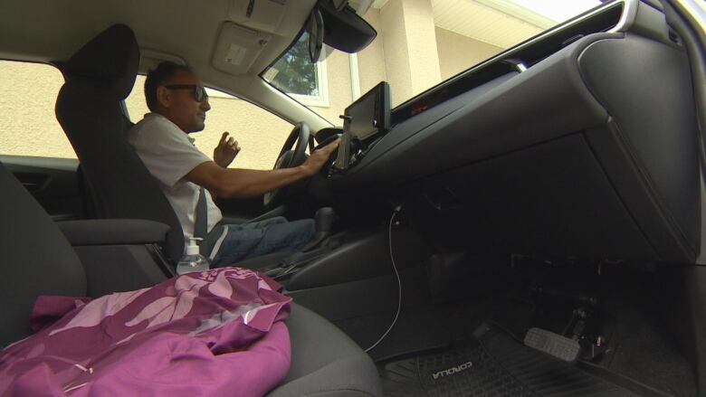 A man in a white shirt wearing sunglasses sits in the driver's seat of his car.