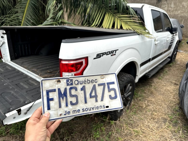 A white 4X4 truck is parked under a tree. A hand holds up a Quebec licence plate found in the bed of the truck.