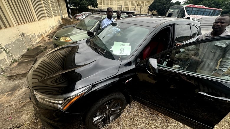 Four men stand beside a black SUV with its doors open in a lot with other cars. 