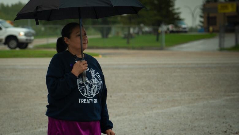 A woman in a Treaty 6 sweatshirt and a skirt walks along holding an umbrella.