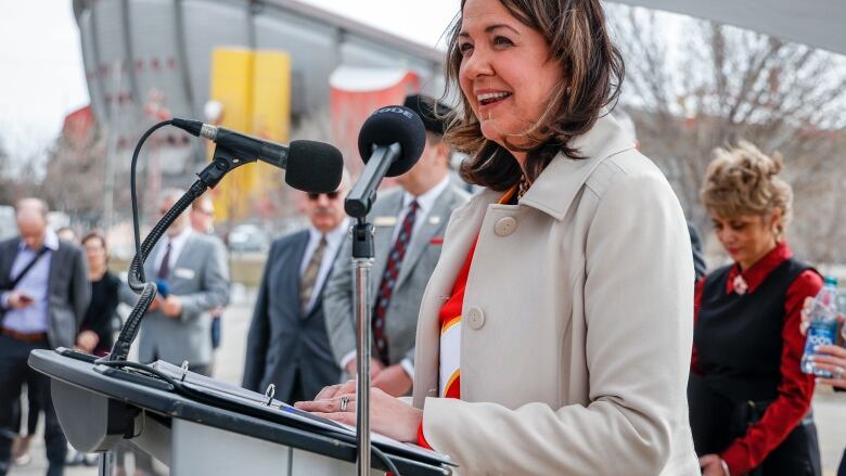 Woman smiles at lectern, wearing a Flames sweater beneath her coat. Other dignitaries and a saddle-shaped hockey arena appear behind her.