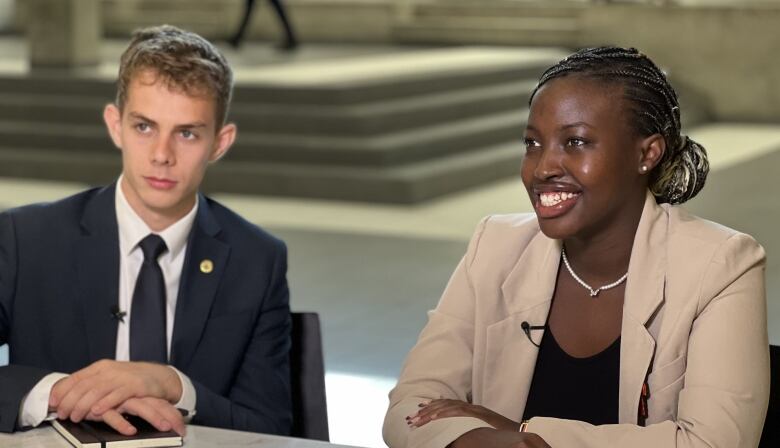 A young woman and man sit at a table smiling, both wearing blazers, as students walk through the corridor behind them.