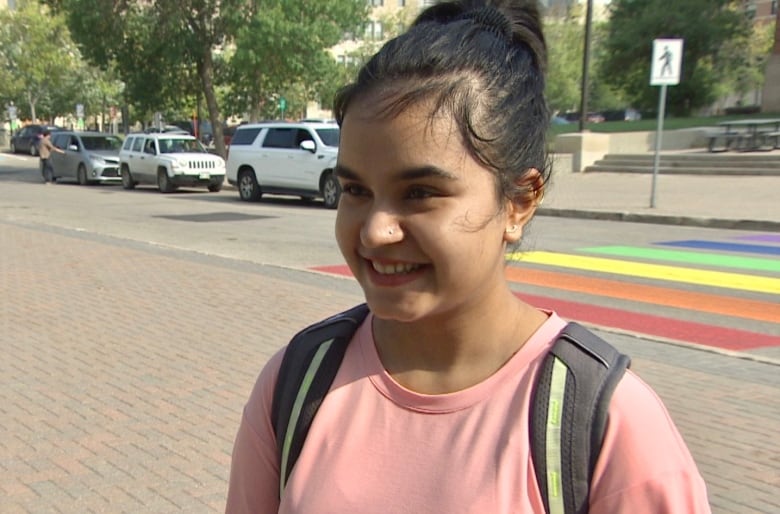 A smiling student wearing a backpack and pink t-shirt stands in front of a rainbow crosswalk on campus.