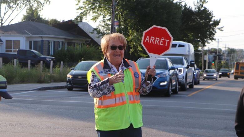 A woman stands in an intersection witha stop sign wearing a vest as a child wearing a backpack walks past 