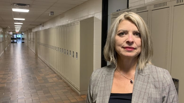 A woman with blond hair in a suit jacket stands in a hallway with school lockers along the walls in the background.